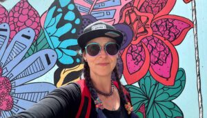A white woman with a black baseball cap, aviator sunglasses, and braids in front of a colorful floral mural by Kelsey Montague.