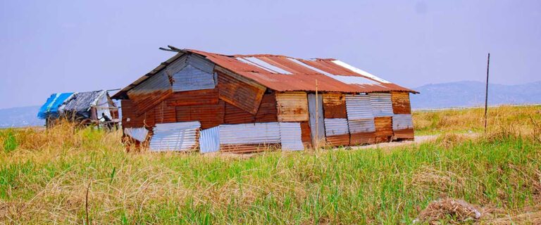 A weathered, patchwork structure built from corrugated metal sheets in varying stages of rust and wear, standing in an open grassy field under a hazy purple-gray sky, with hills visible in the distance. (Alt text generated by Claude and human-reviewed)
